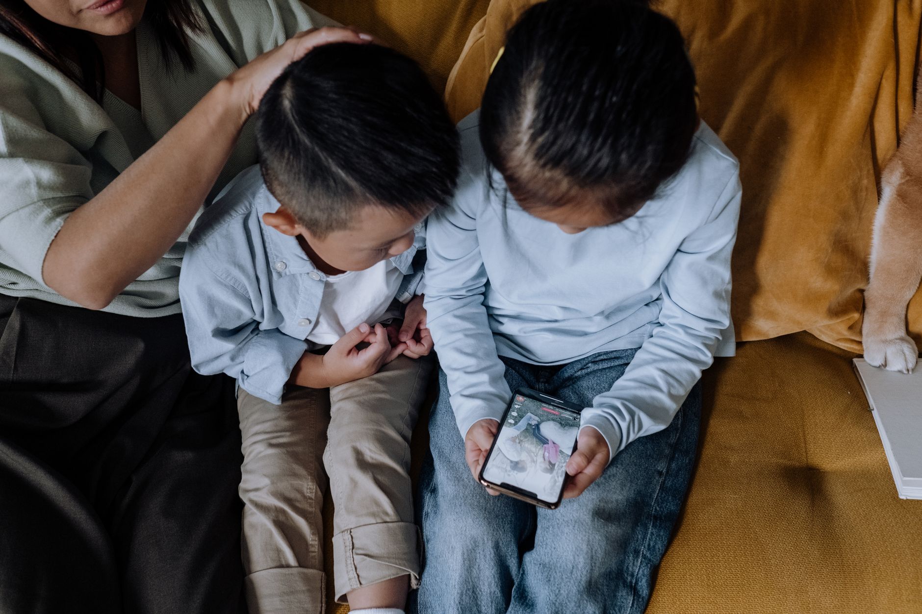 Deux enfants assis sur un canapé jaune regardent un smartphone, tandis qu’un adulte pose la main sur la tête de l’un d’eux.