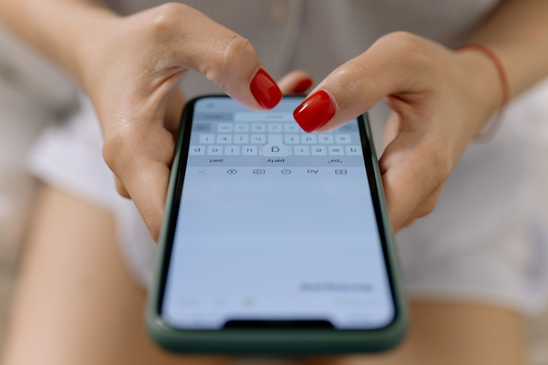 Deux mains aux ongles rouges tapent sur le clavier d’un smartphone à coque verte, tenu au-dessus des genoux.